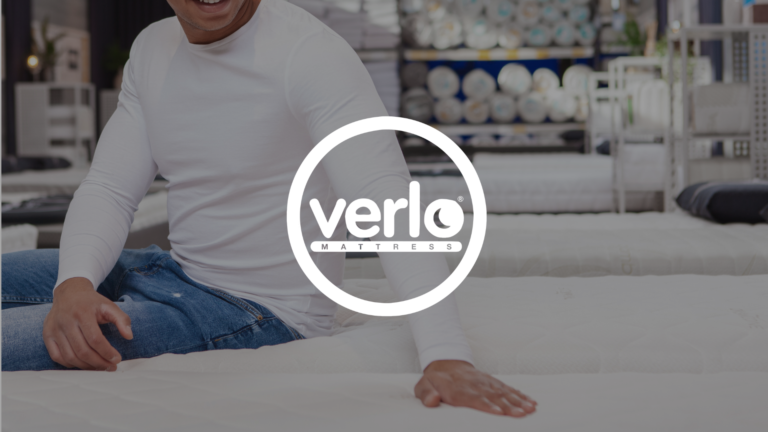 A tan man wearing a long sleeved white shirt and jeans sits on the edge of a mattress inside of a mattress store with an outstretched hand testing the mattress next to him, his face is out of the photo. A white Verlo Mattress logo overlays the photo.