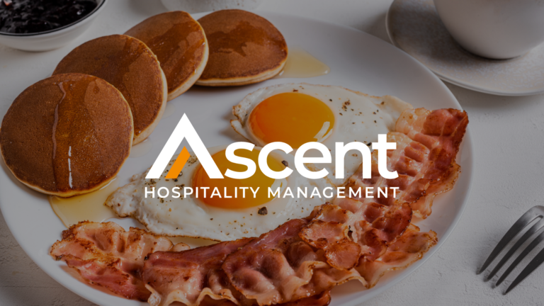 A white plate of bacon, two eggs over easy, and four pancakes with syrup are shown behind a white Ascent HM logo.